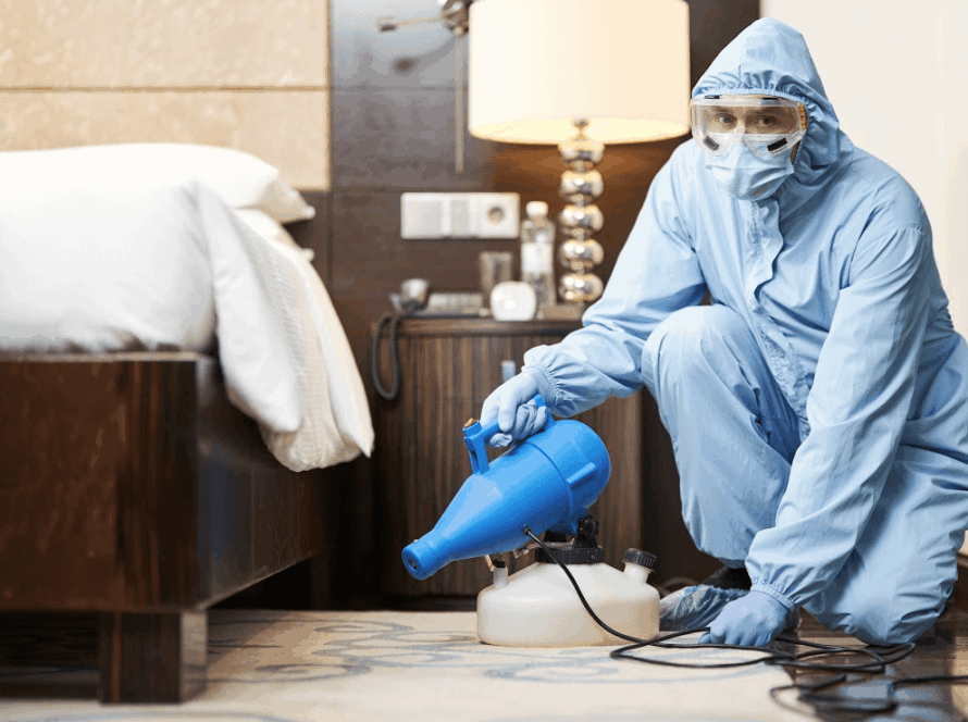 A pest control worker in a biohazard suit is disinfecting underneath a bed.